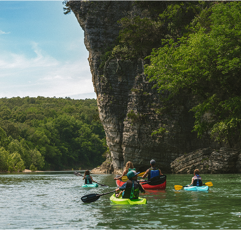 Kayakers paddle near a rocky cliff on a calm river surrounded by lush trees.