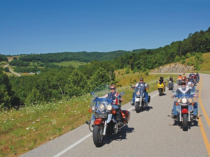 Motorcyclists ride on a scenic rural road under a clear blue sky.