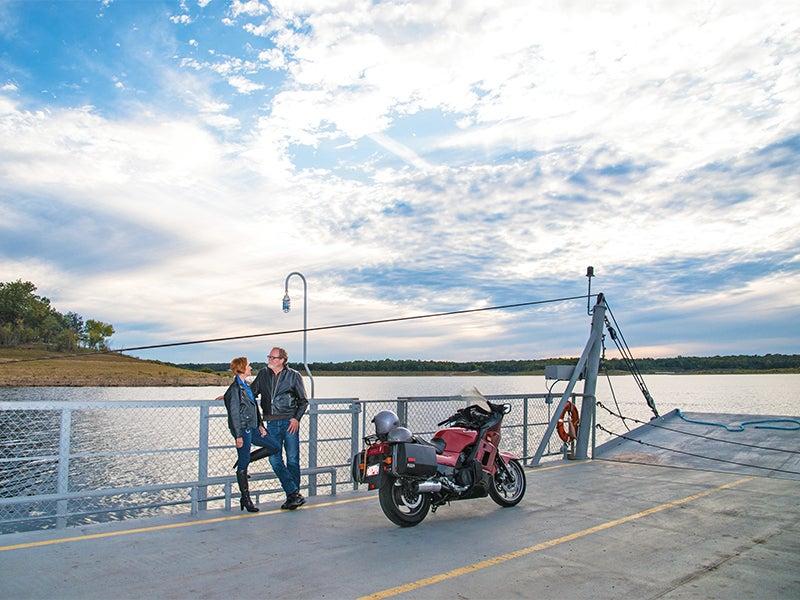 Two people and a motorcycle on a ferry, with a cloudy sky and water background.