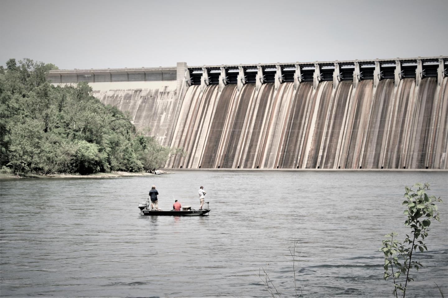 Boat with people on a lake near a large dam, surrounded by trees.