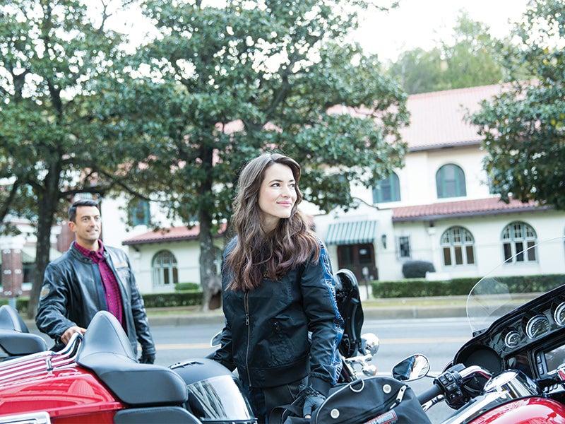 Two people in front of motorcycles on a tree-lined street, with a building in the background.