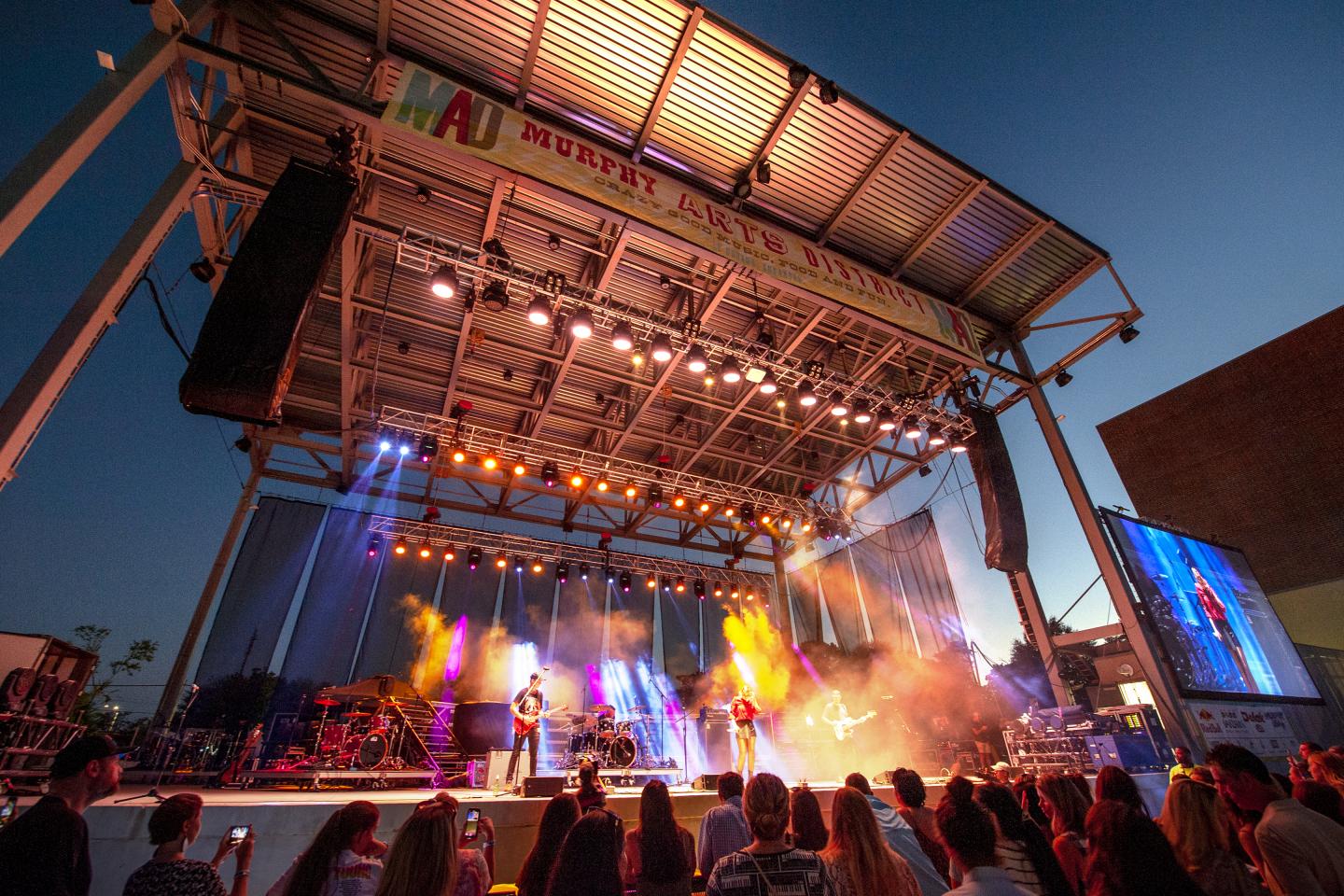 Concert stage with colorful lights and performers, crowd watching at sunset.