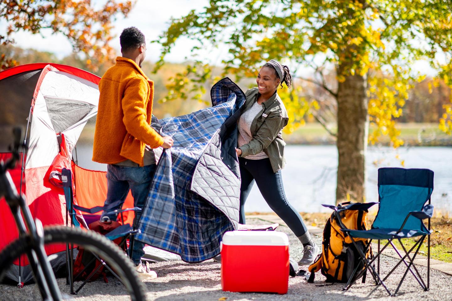 Couple setting up a tent by a lake; autumn trees in the background.