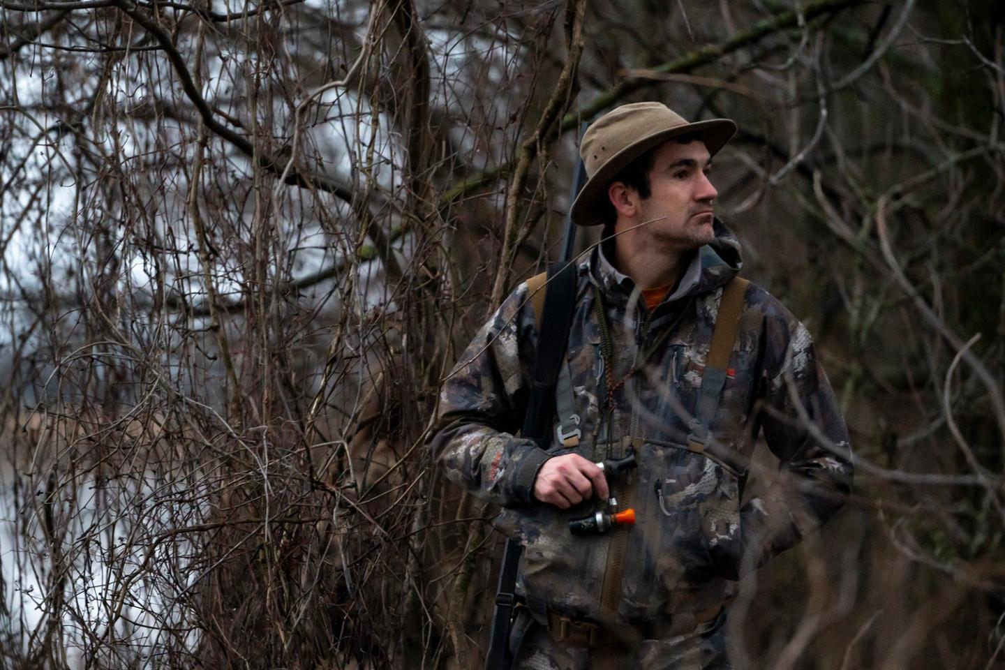 Hunter in camouflage gear stands in a wooded area, holding a camera.