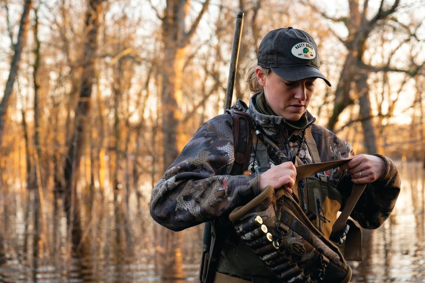 Hunter in camouflage adjusts gear in flooded forest.
