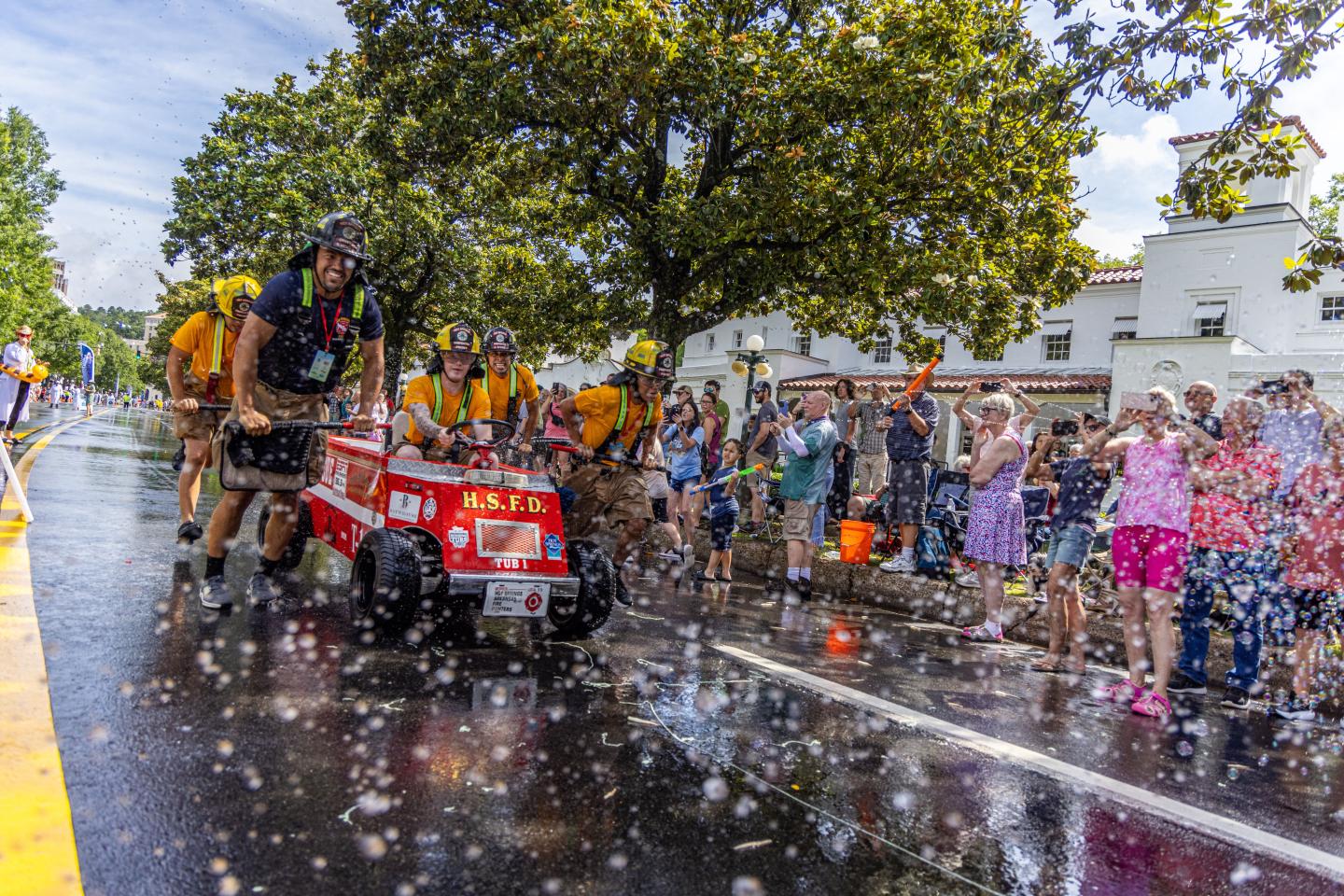 Parade with red cart and costumed participants, surrounded by bubbles and spectators.