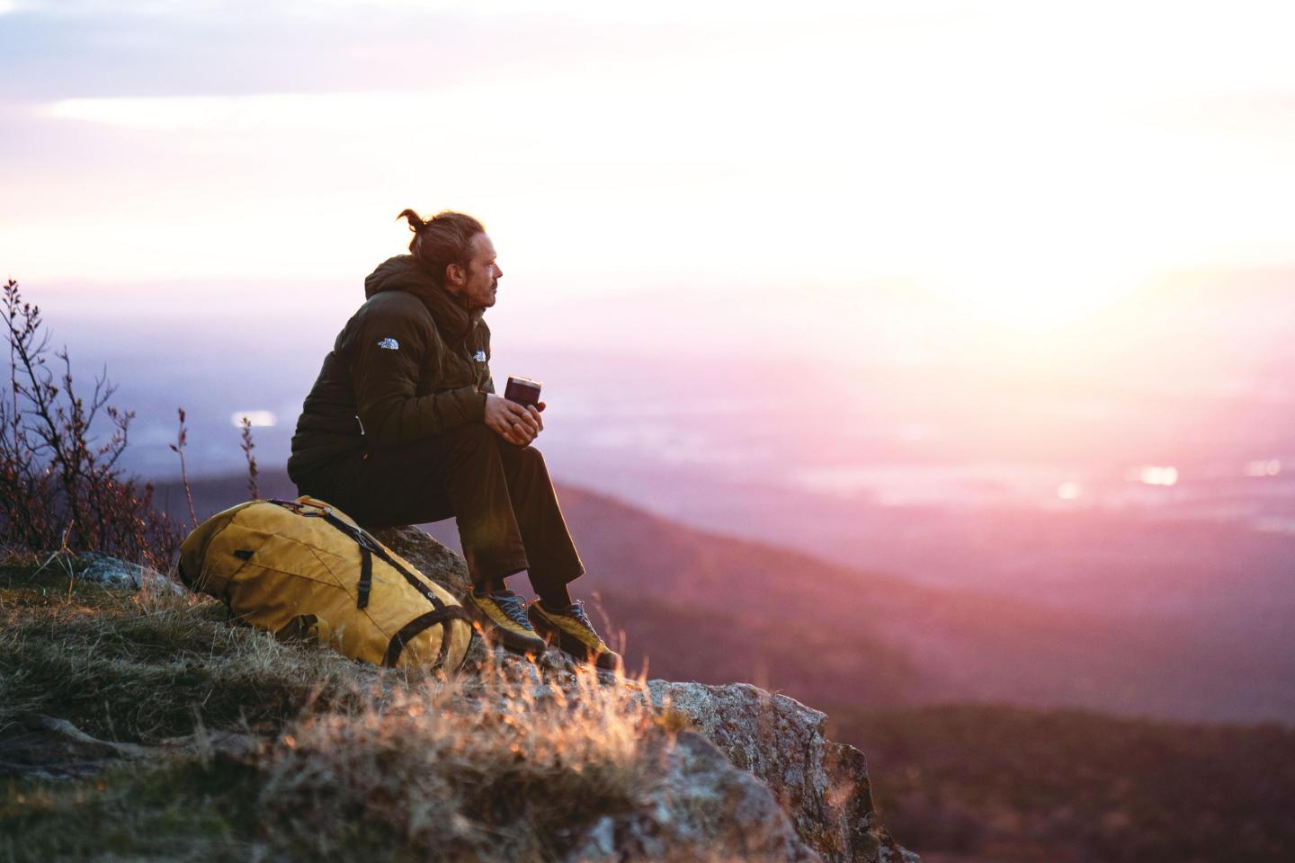 Man sitting on a mountain, holding a mug at sunrise; yellow backpack beside him.