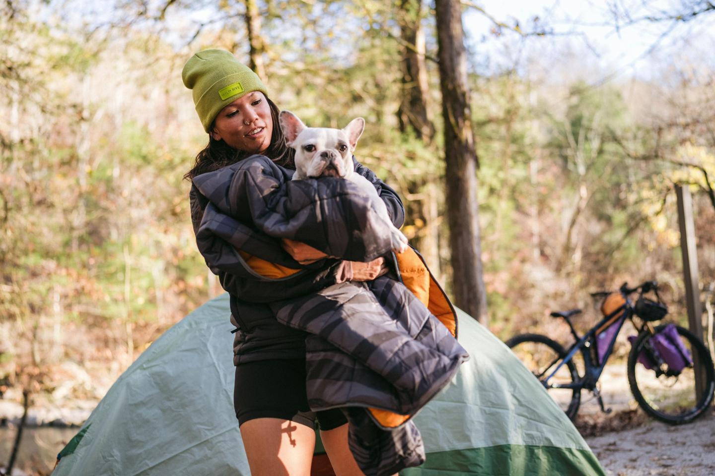 Woman holding a dog, standing by a tent in a wooded area.