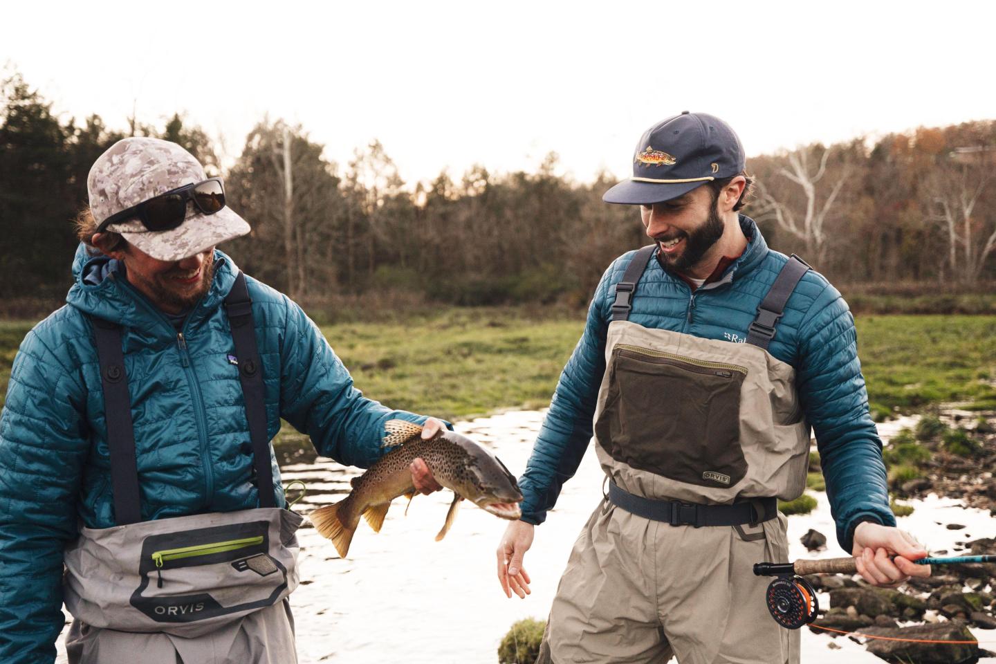Two men in fishing gear holding a fish by a river.