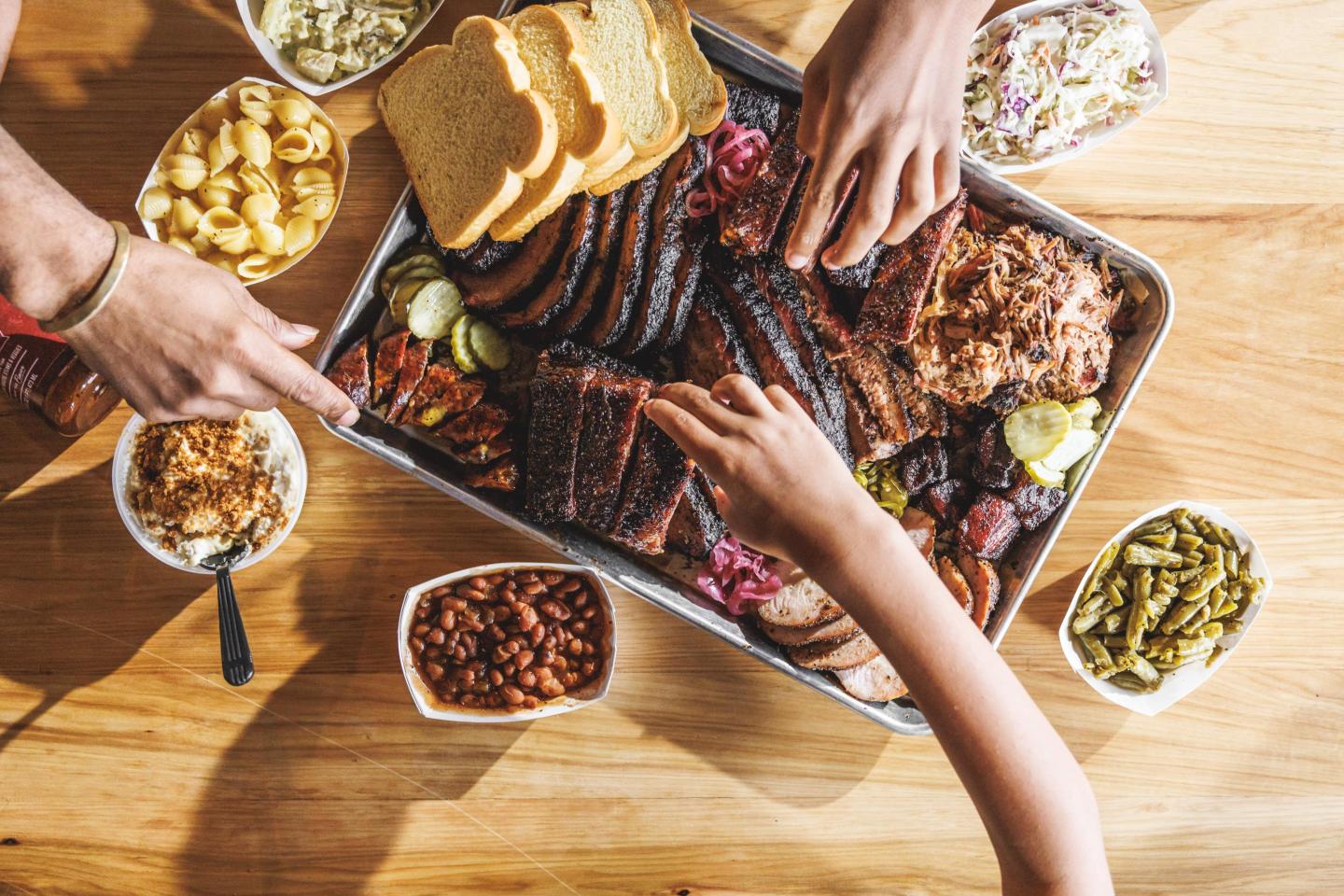Hands reaching for barbecue meats on a large platter with sides.