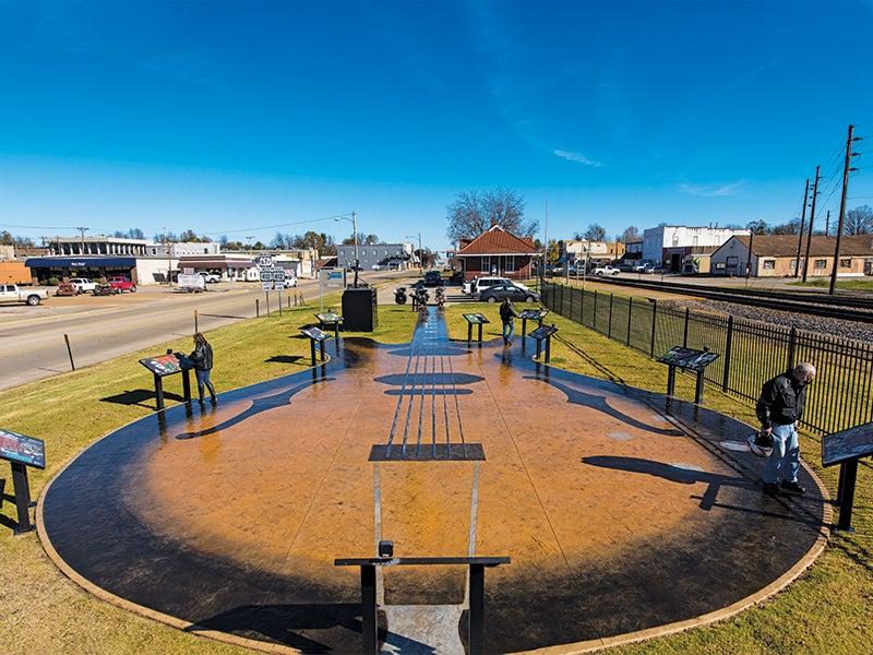 Guitar-shaped plaza with signs, surrounded by grass and a fence under a clear blue sky.