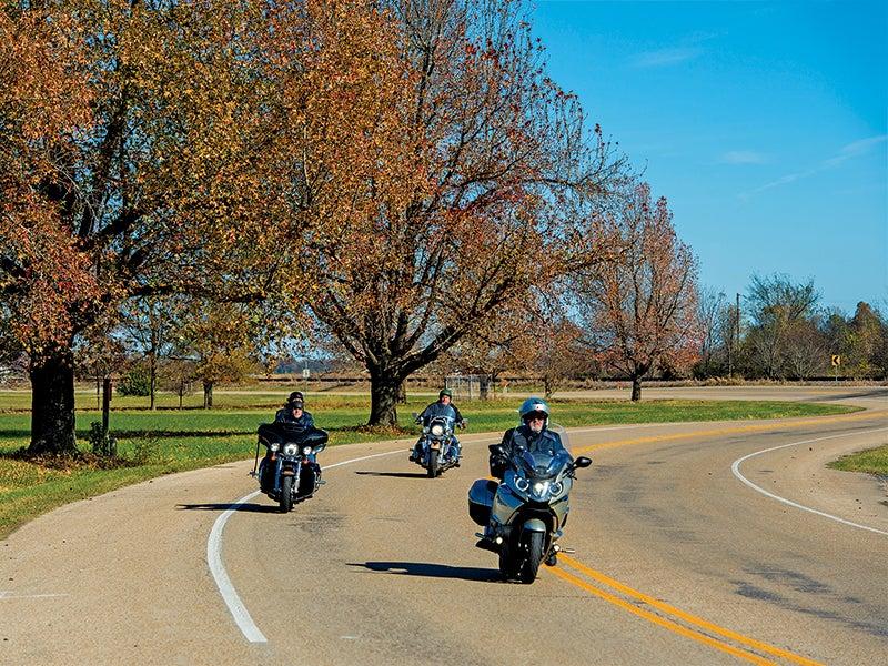 Motorcyclists ride on a winding road lined with autumn trees under a blue sky.