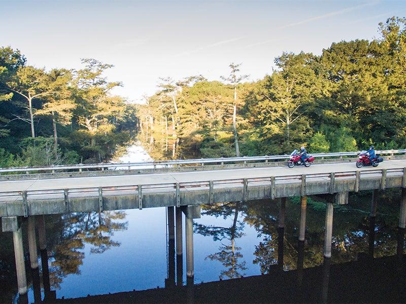 Motorcycles cross a bridge over a calm river, surrounded by trees.
