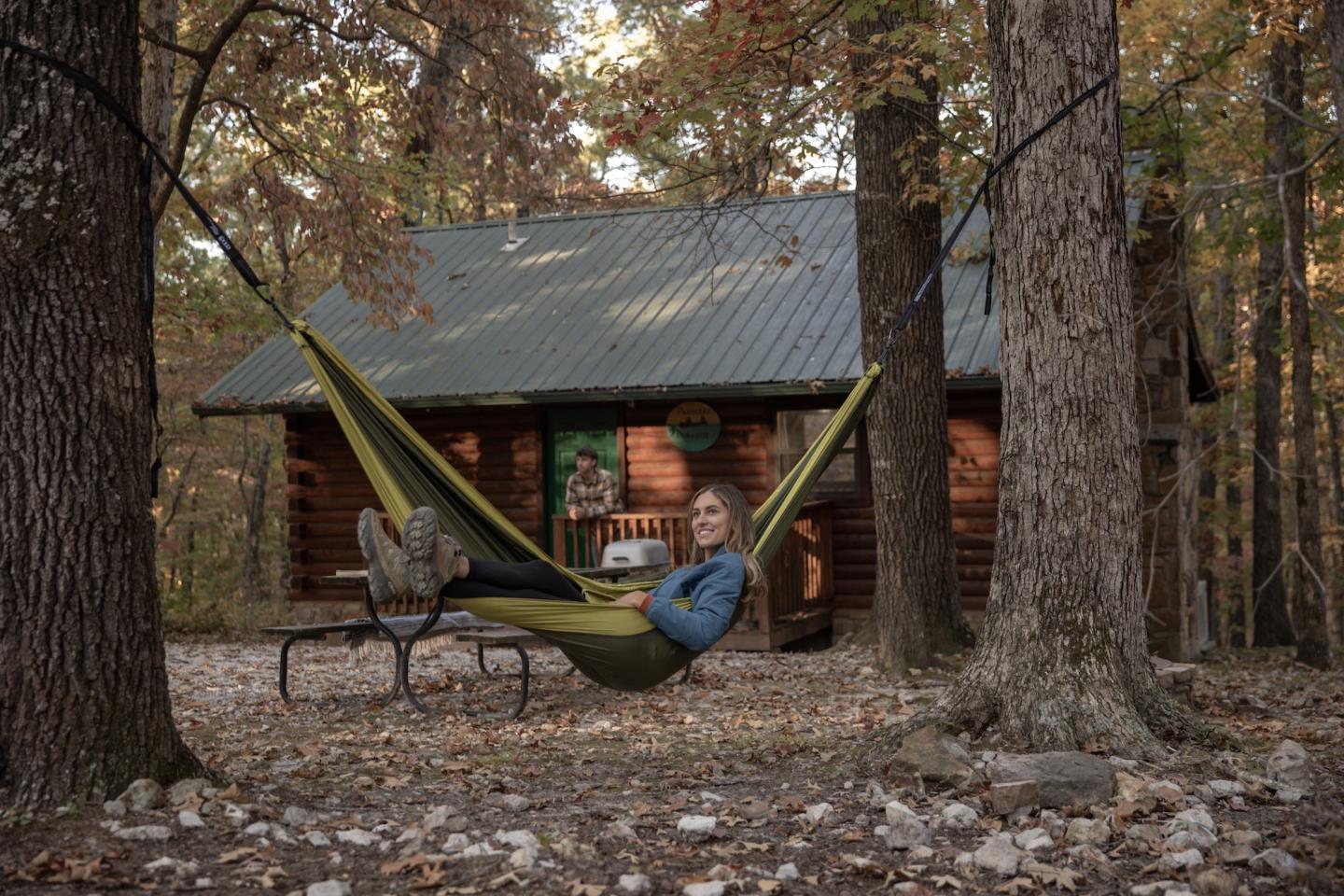 Person relaxing in a hammock between trees near a cabin in autumn woods.