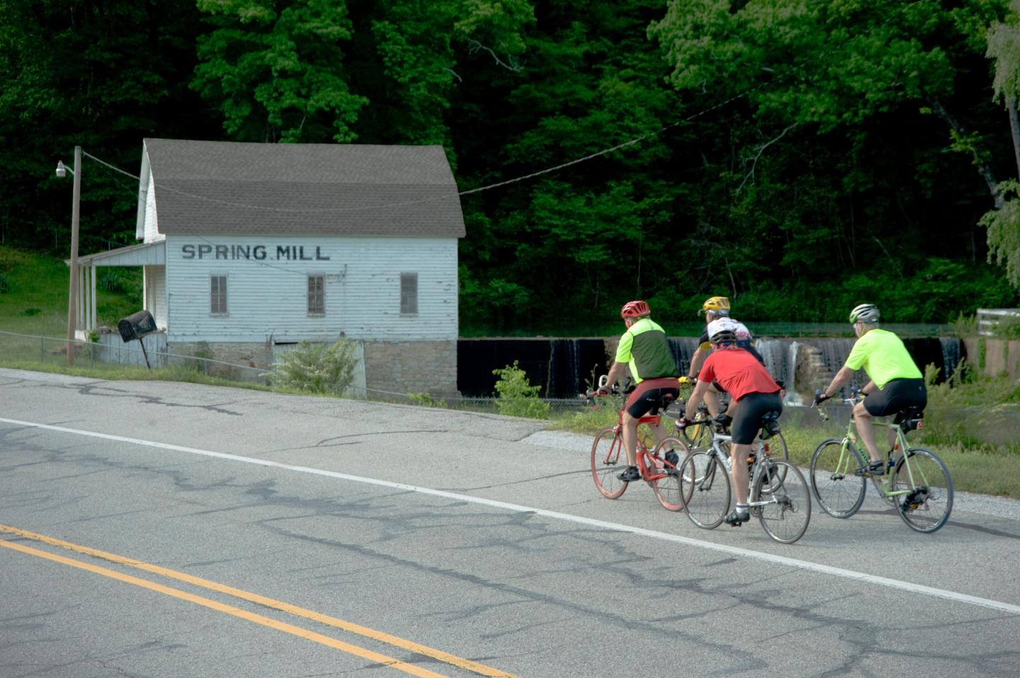 Cyclists ride past a small white building near a lush green area.