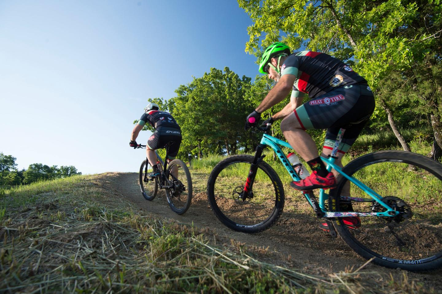 Cyclists riding uphill on a dirt trail, surrounded by trees and blue sky.