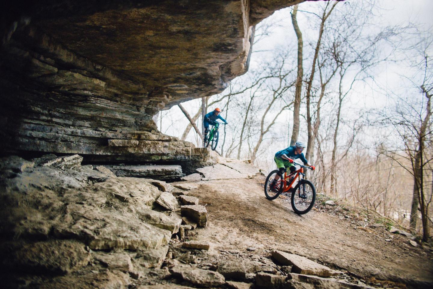 Mountain bikers ride along a rocky forest trail under a stone overhang.