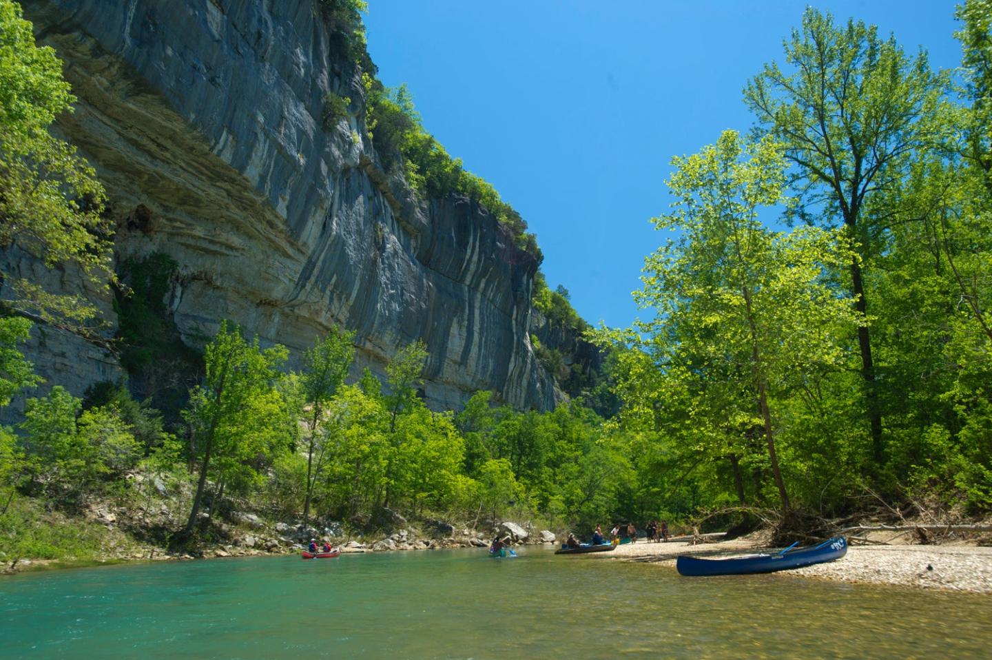 Clear river with canoes, trees, and a rocky cliff under a bright blue sky.