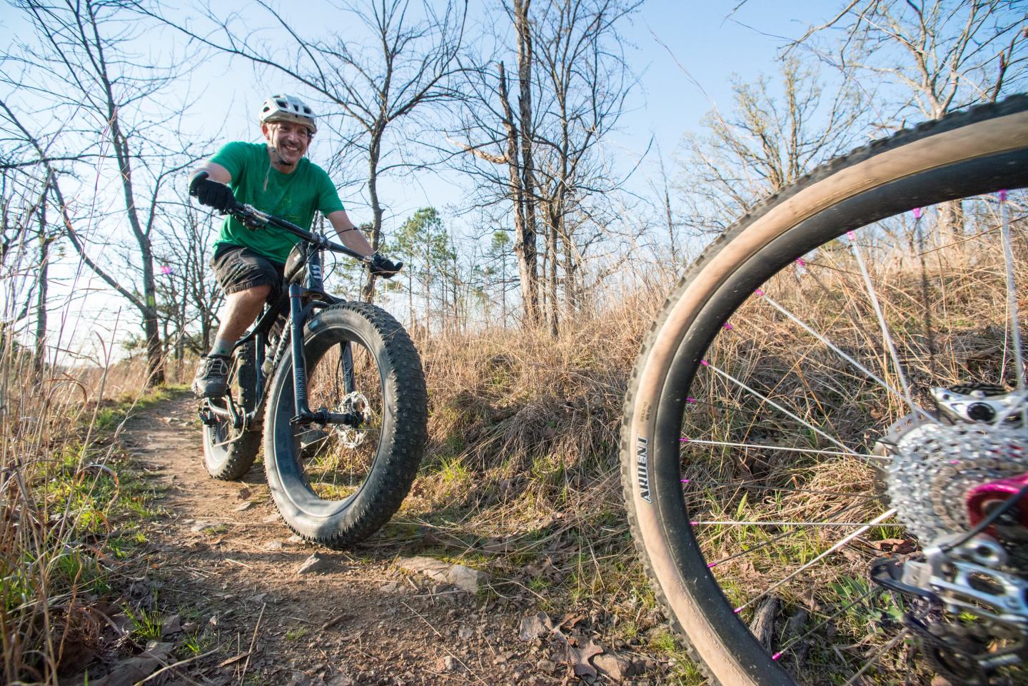 Man biking on a dirt trail, surrounded by bare trees, sunny day.