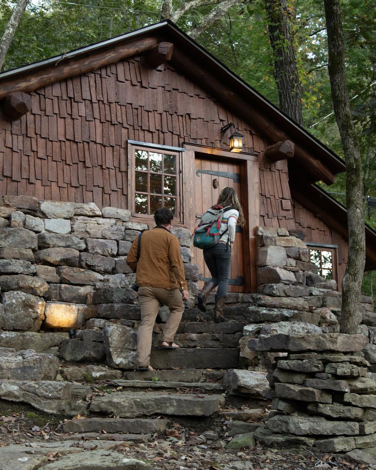 Cozy cabin in the woods, two people approaching the door on stone steps.