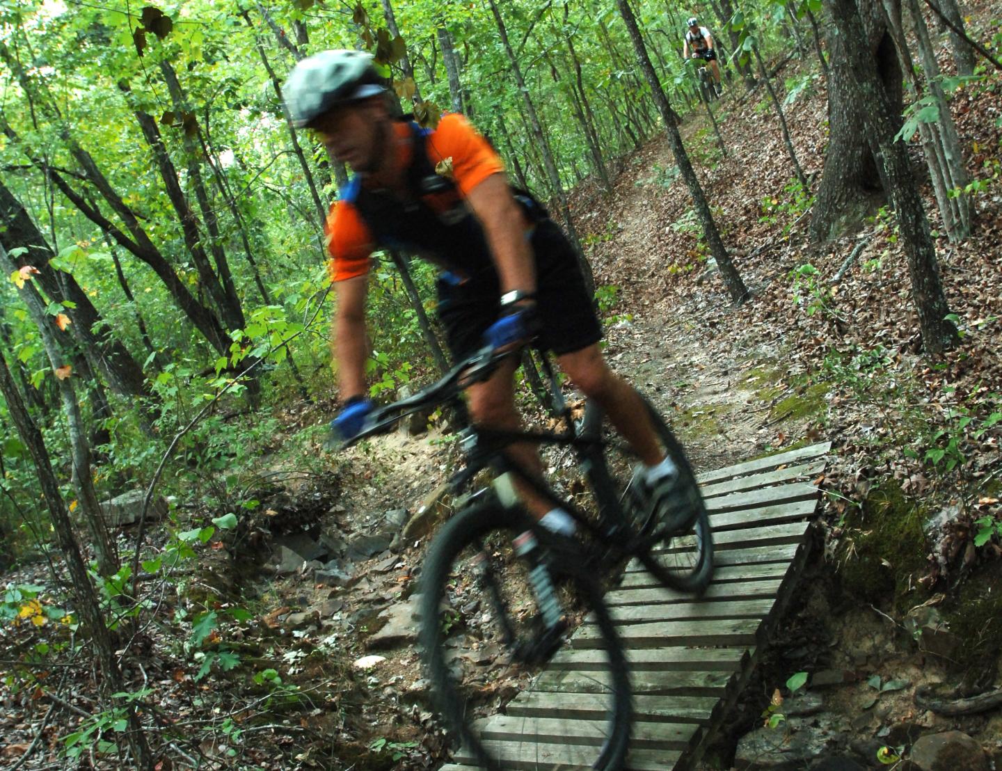 Cyclist on a forest trail bridge, wearing a helmet and an orange shirt.