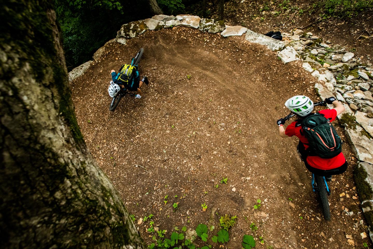 Two mountain bikers navigate a dirt trail curve in a wooded area.