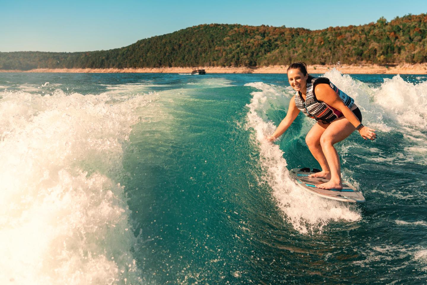 Woman wakeboarding on a sunny lake, surrounded by waves, wearing a life vest.