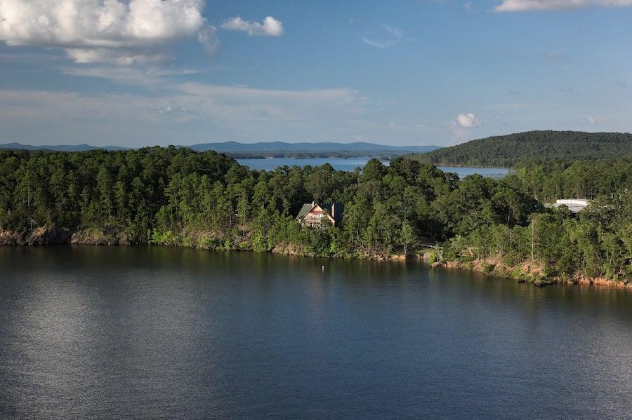 Calm lake with forested shore, single house, and distant hills under blue sky.