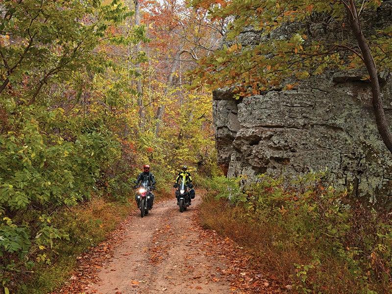 Two motorcyclists on a dirt path through a colorful autumn forest.