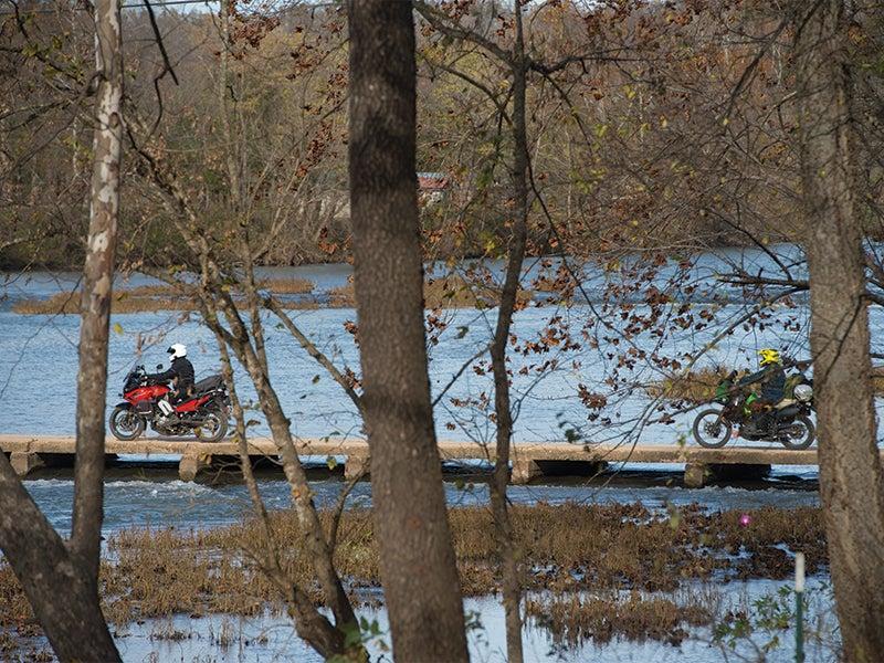 Motorcycles crossing a wooden bridge over a calm river, surrounded by autumn trees.