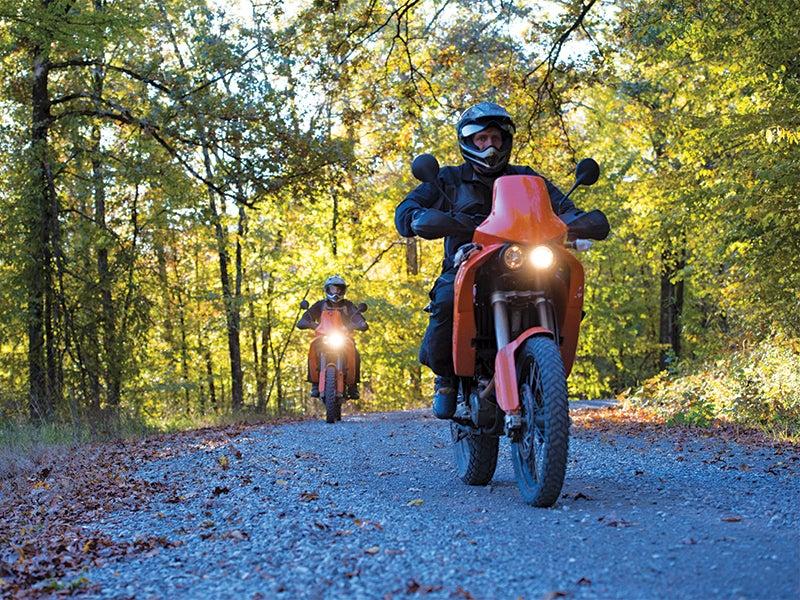 Two motorcyclists on a forest trail in autumn, sunlight filtering through trees.