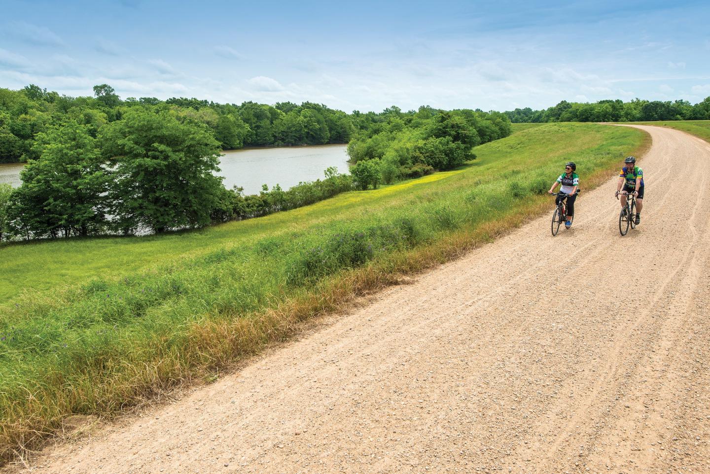 Two cyclists on a gravel road beside a river and green fields.