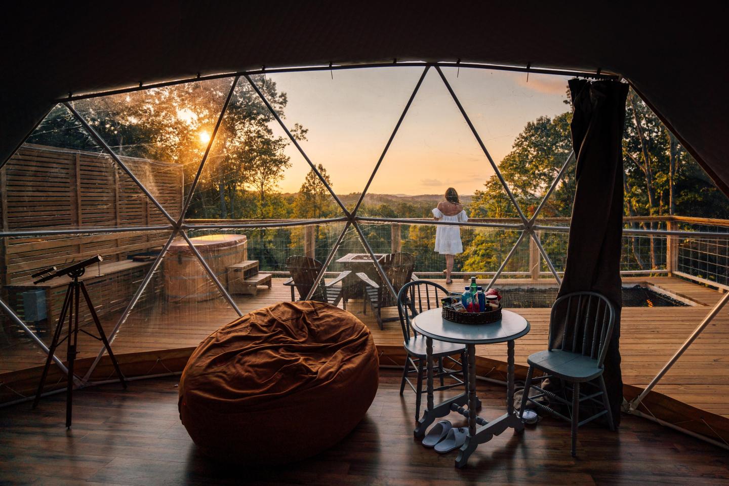 Geodesic dome interior with a view of sunset, woman on balcony, bean bag, and table with chairs.