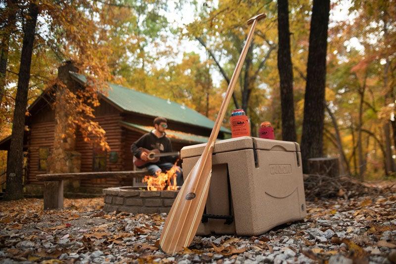 Cabin in autumn woods, person by fire, cooler and paddles nearby.