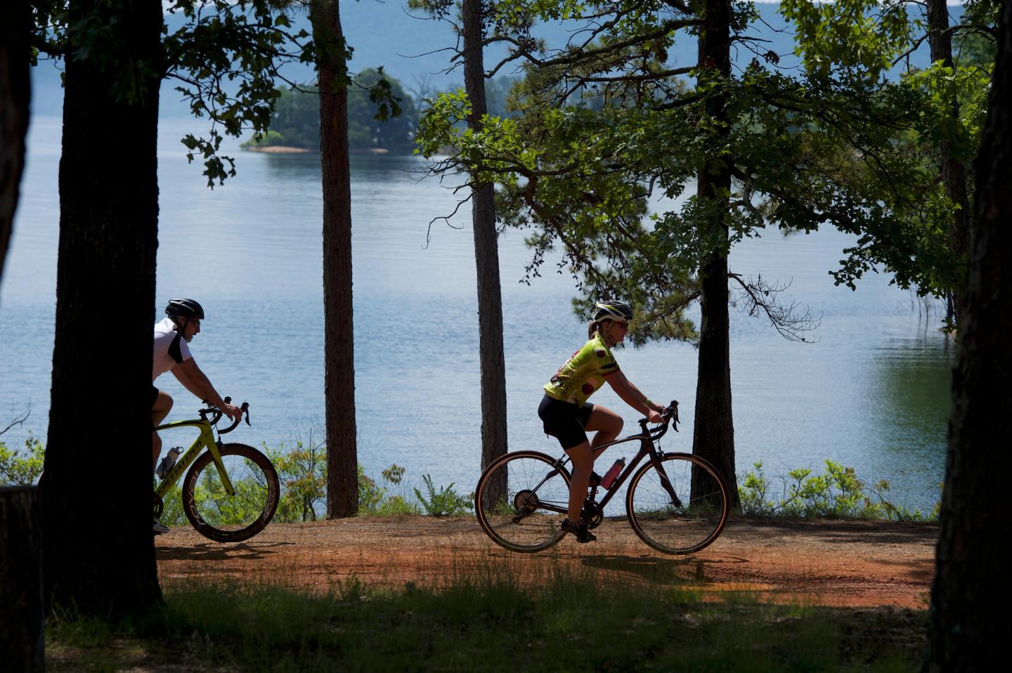 Cyclists ride on a forest trail beside a calm lake, under tall trees.