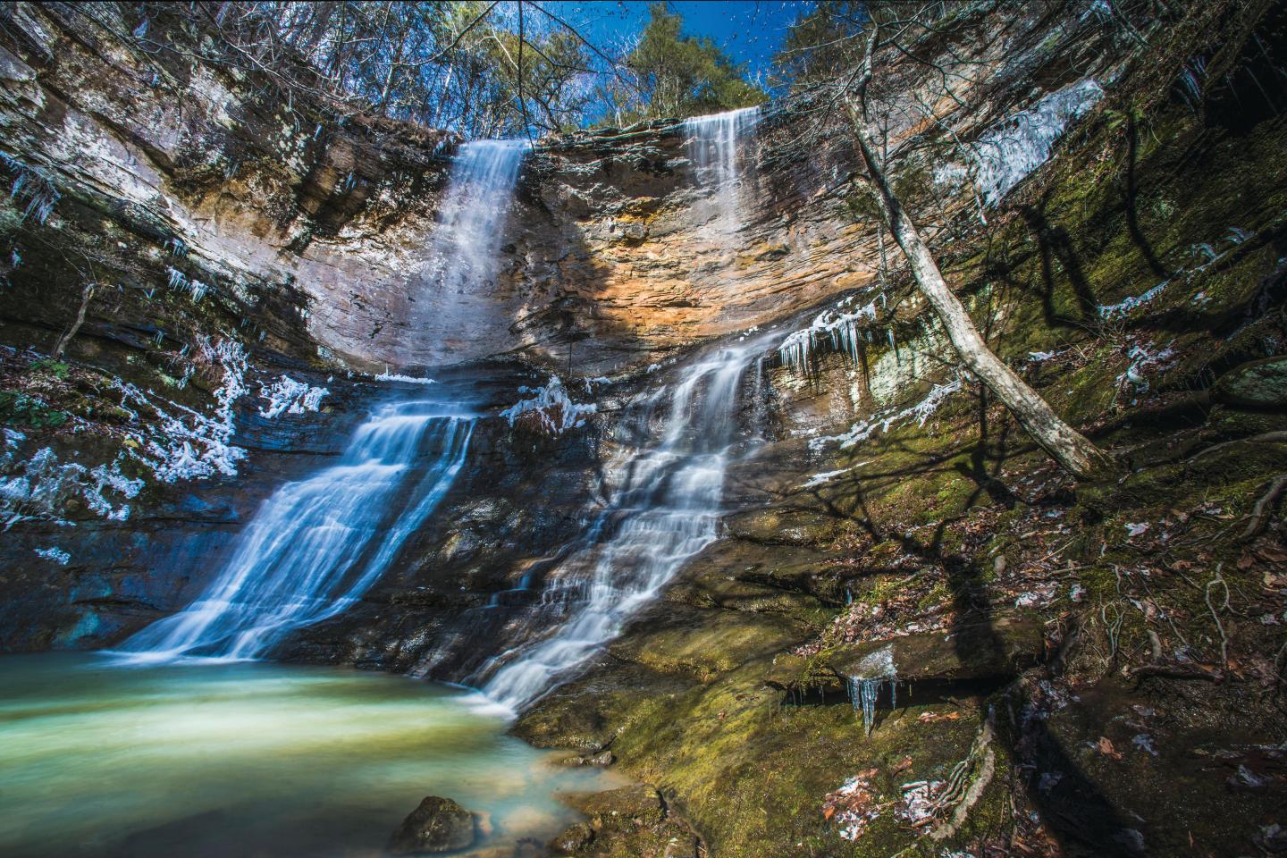 Two cascading waterfalls in a forest with sunlight and mossy rocks.