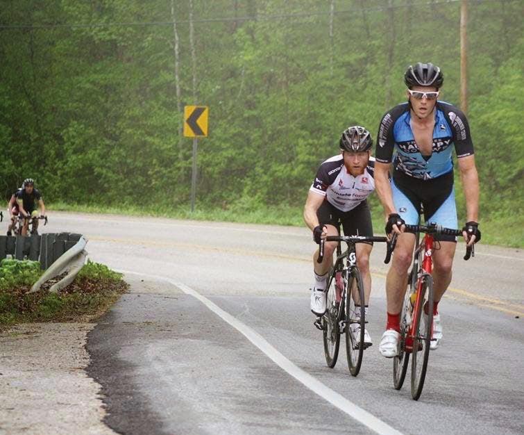 Cyclists riding uphill on a forested road, with overcast skies.