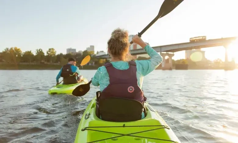 Two people kayaking on a river beneath a sunny bridge.