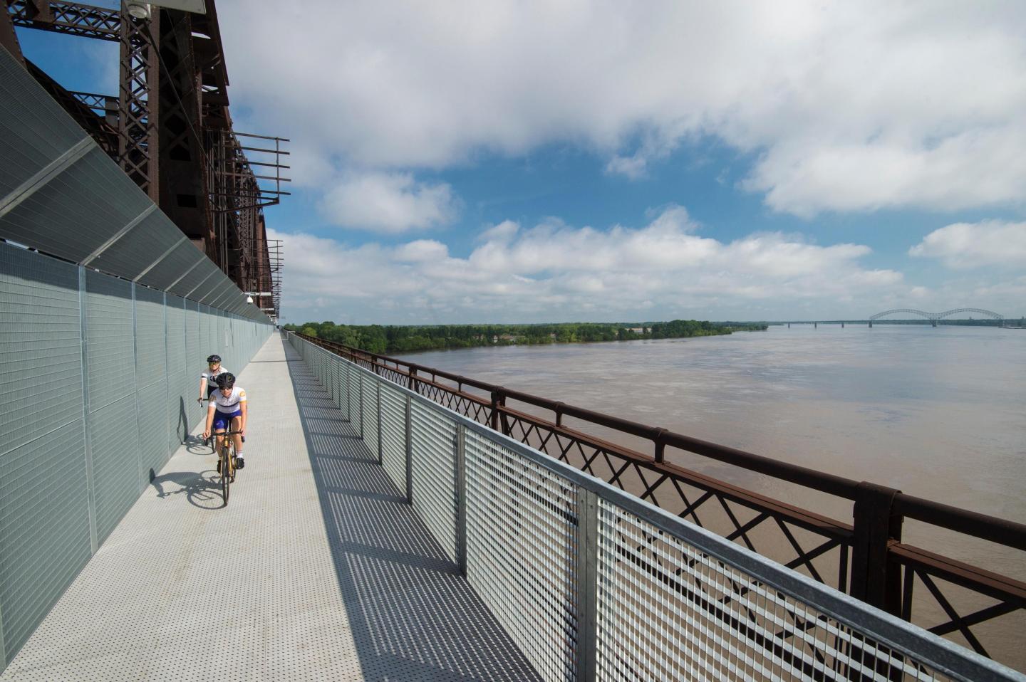 Person biking on a bridge with river view under a cloudy sky.