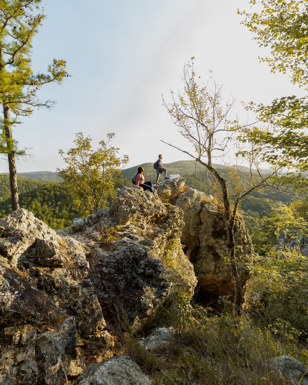Two people sitting on a rocky cliff surrounded by trees and hills.