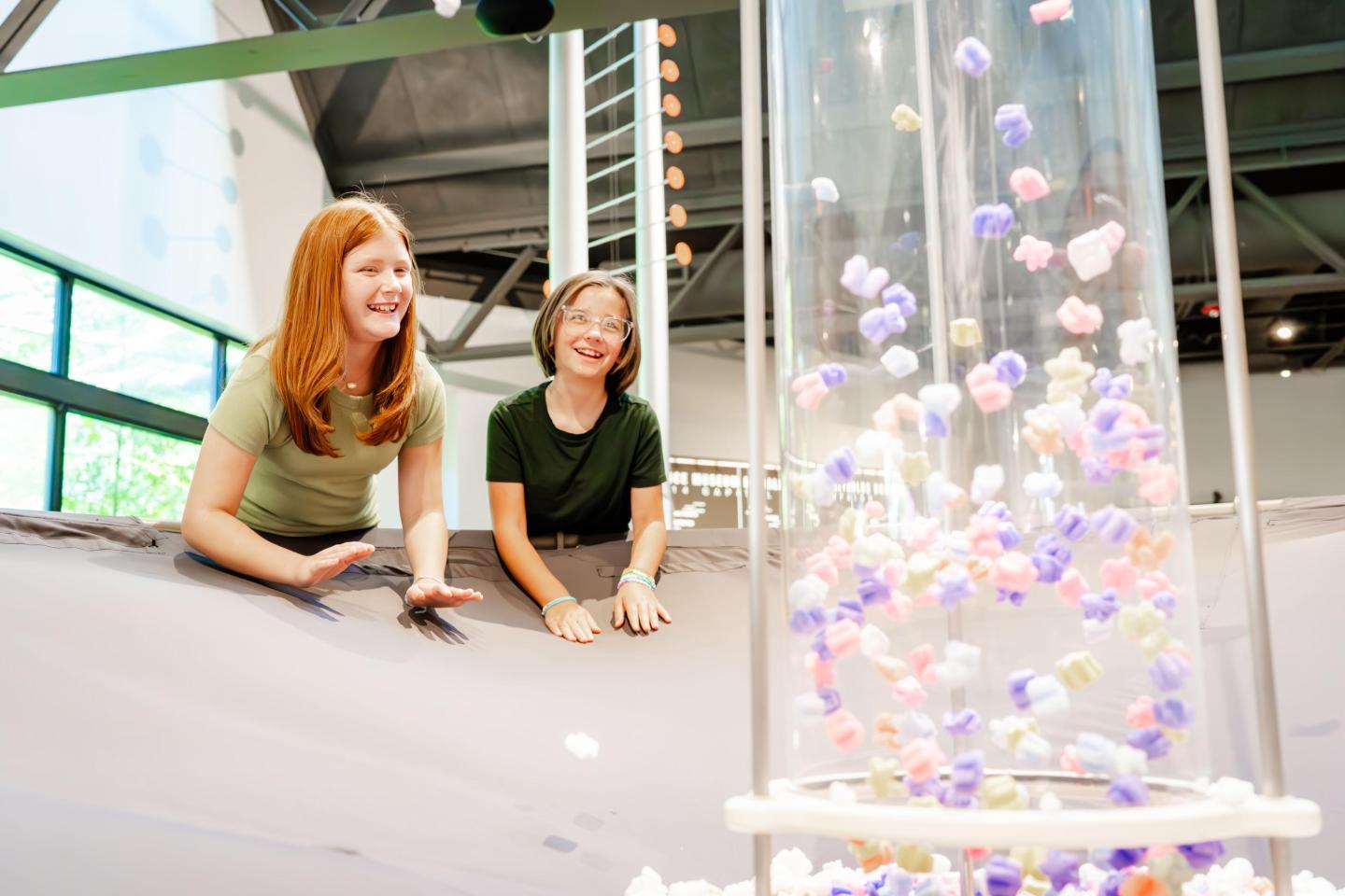 Kids laughing near a tube of colorful balls indoors.
