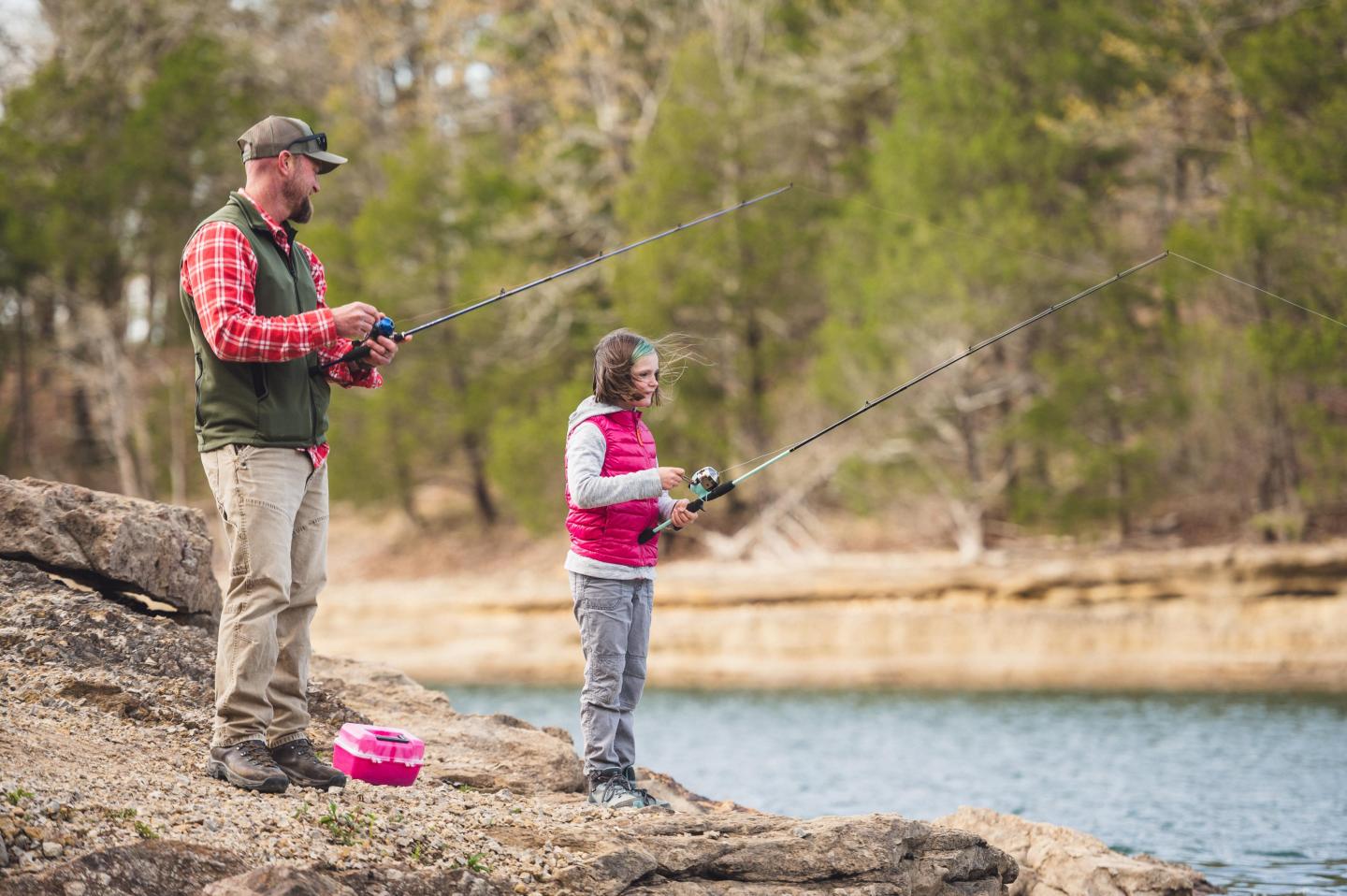 Man and child fishing at a lakeside, surrounded by trees.