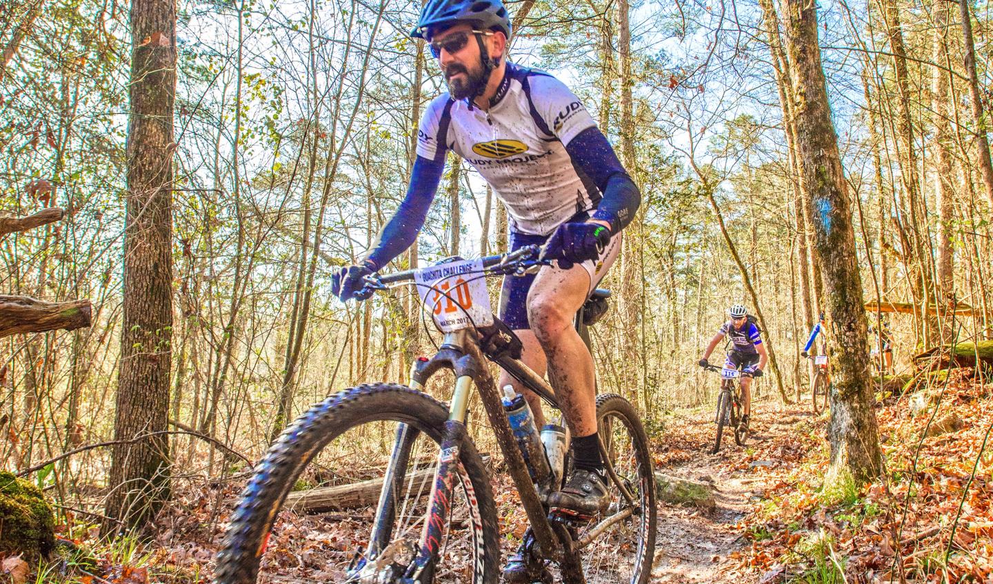 Mountain biker riding through a forest trail, wearing blue and white gear.