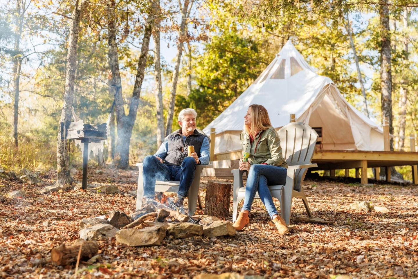 Two people sit by a campfire, with a white tent and trees in the background.