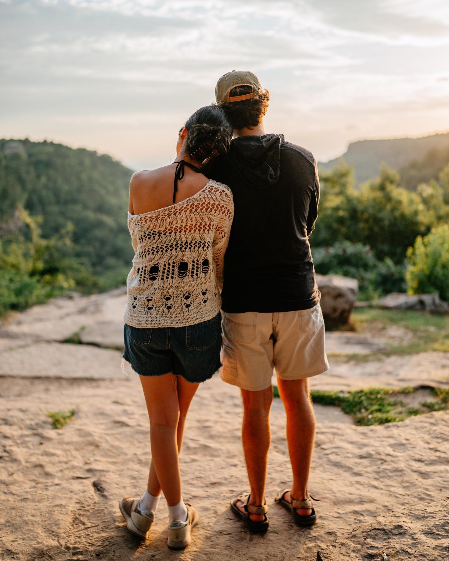 Couple standing on a rocky path, overlooking a scenic, tree-filled landscape at sunset