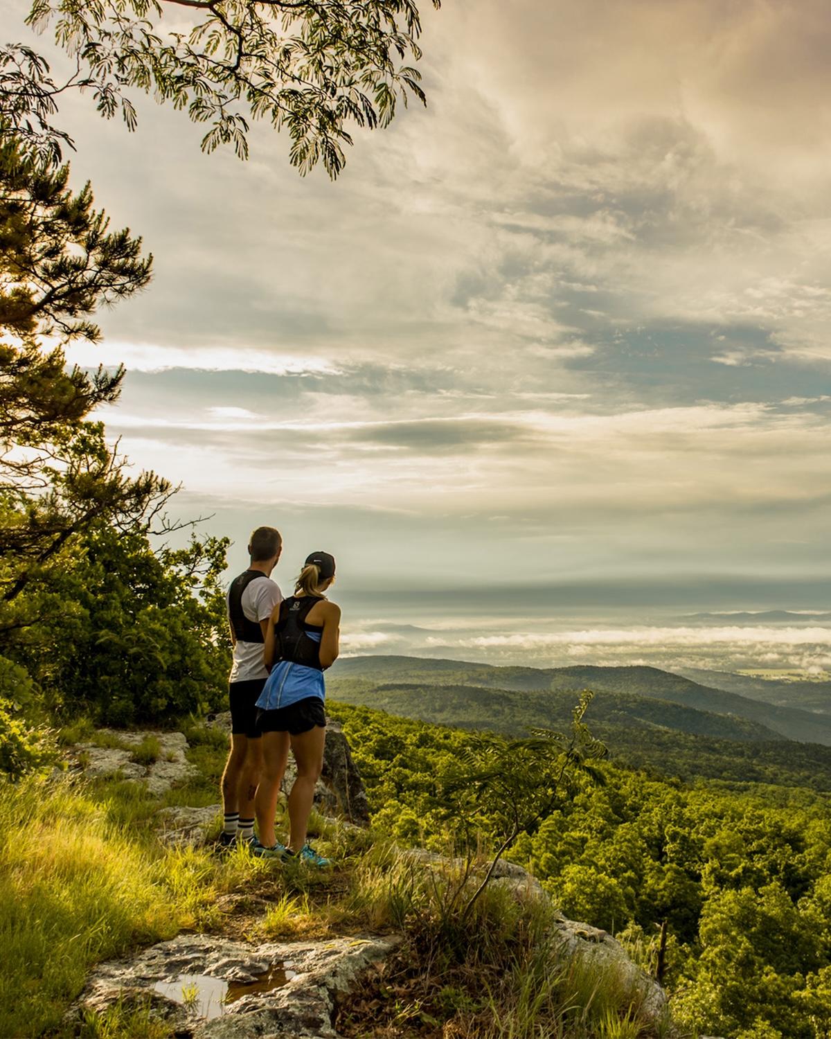 Two people in athletic gear admire a scenic mountain view under a cloudy sky.