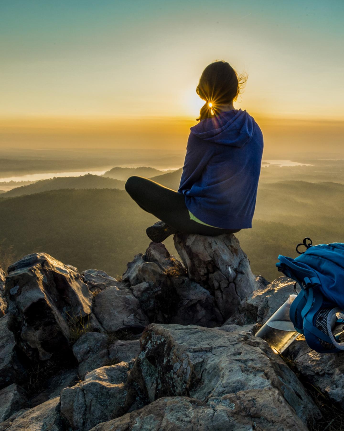 Person sitting on rocky peak, watching a sunset over distant hills.