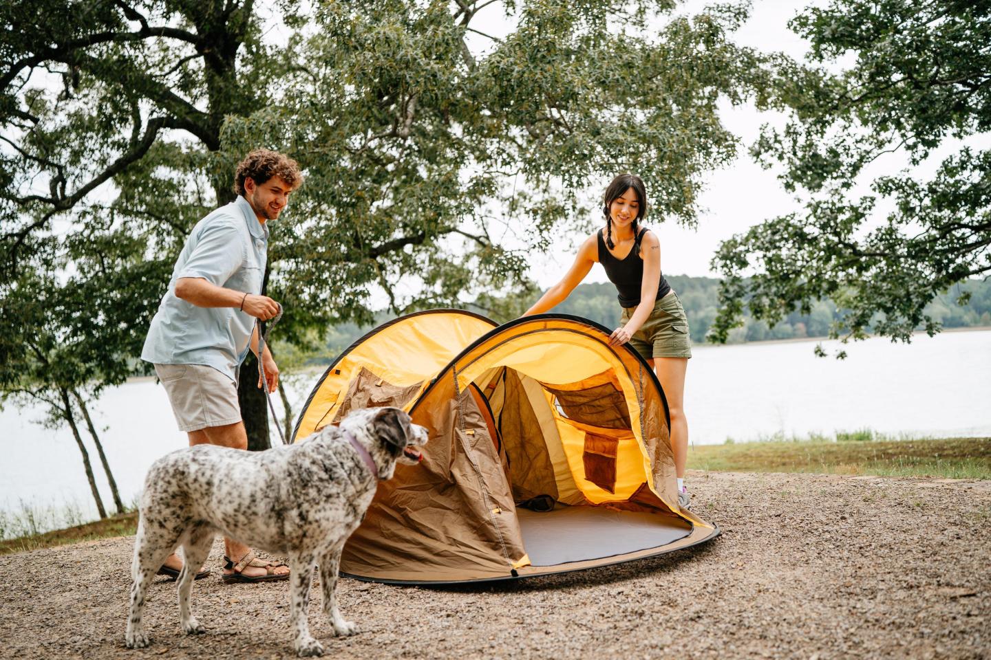 Two people setting up a yellow tent by a lake, with a dog nearby.