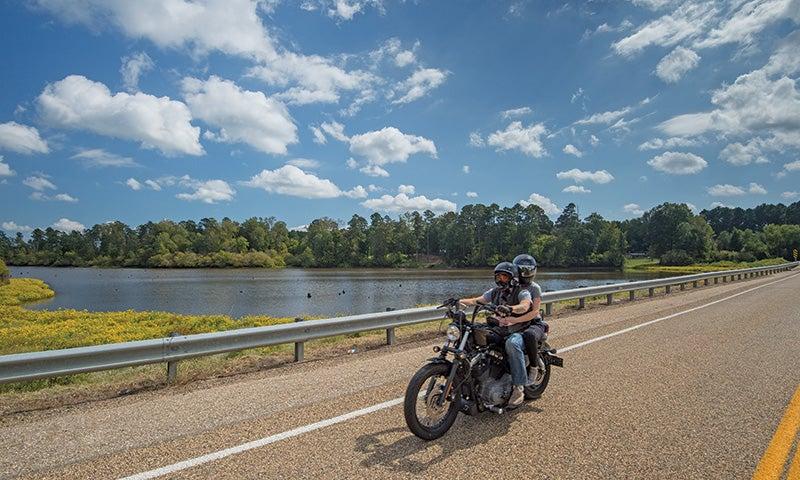 Motorcyclist rides on a sunny road by a lake under a blue sky with clouds.