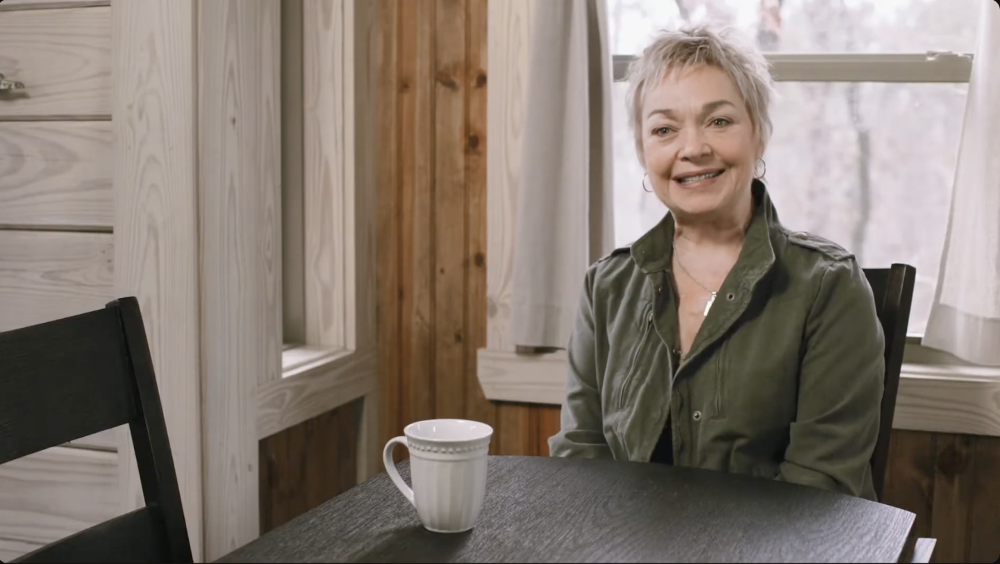 Smiling woman sitting at a table with a mug, wooden wall in the background.